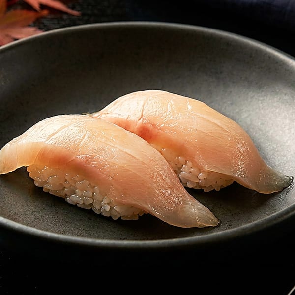 Two pieces of light-colored fish nigiri sushi on a dark plate with chopsticks and autumn leaves.