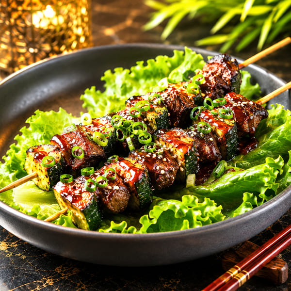 Grilled beef and zucchini skewers with teriyaki glaze, sesame seeds, and green onions on lettuce.